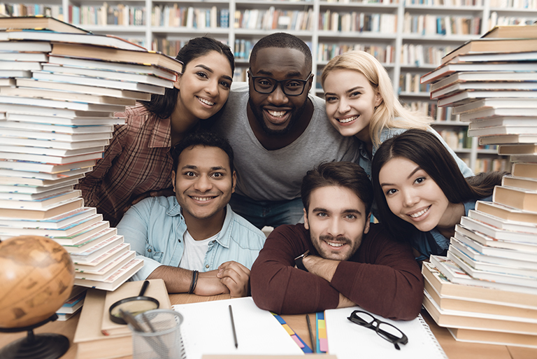 Six ethnic students, mixed race, indian, asian, african american and white sitting at table surrounded with books at library.